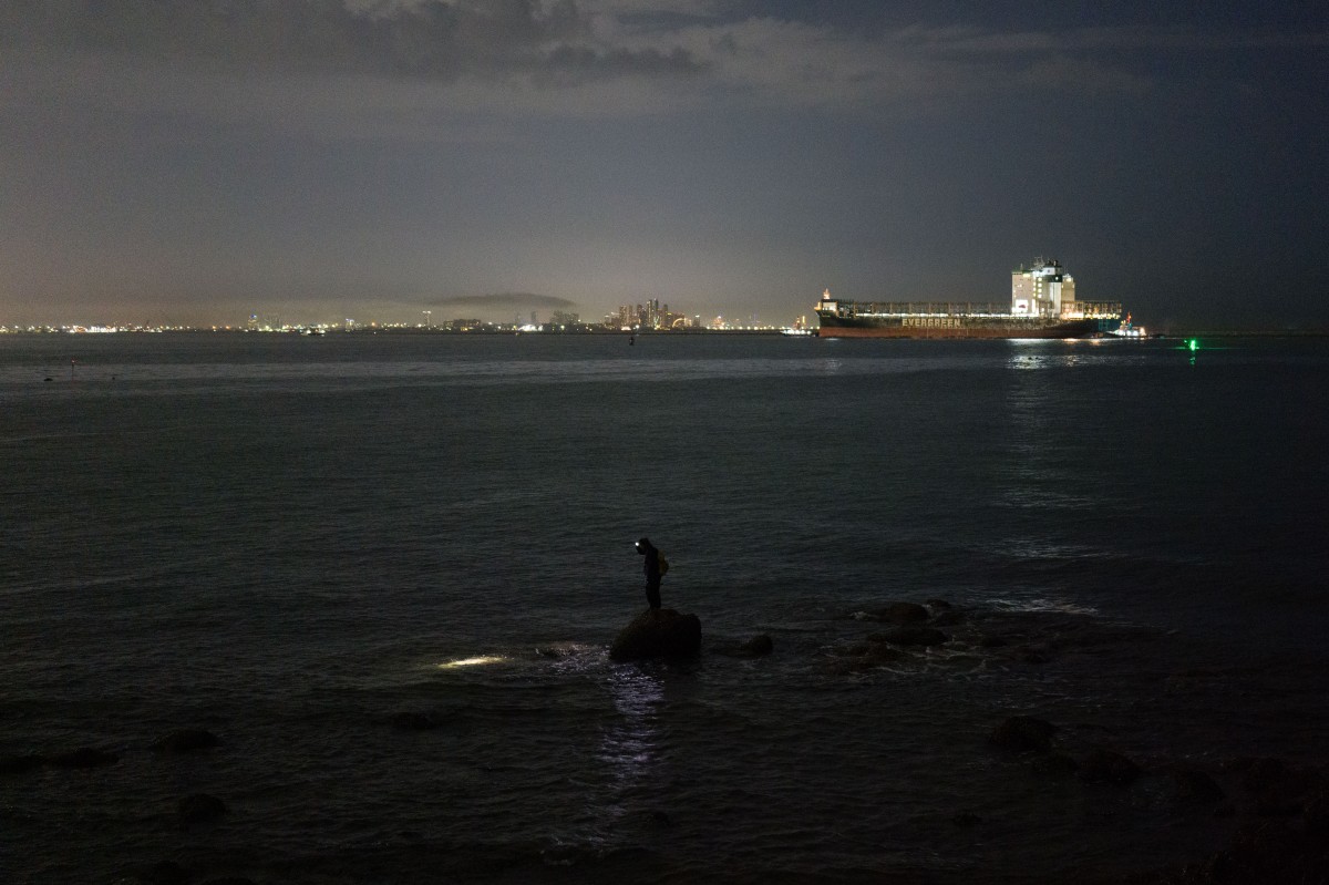 An angler (C) uses a headlamp before casting his line, as a large cargo container ship sails towards the Laem Chabang Port, near the city of Pattaya on September 1, 2026. (Photo by ANTHONY WALLACE / AFP)