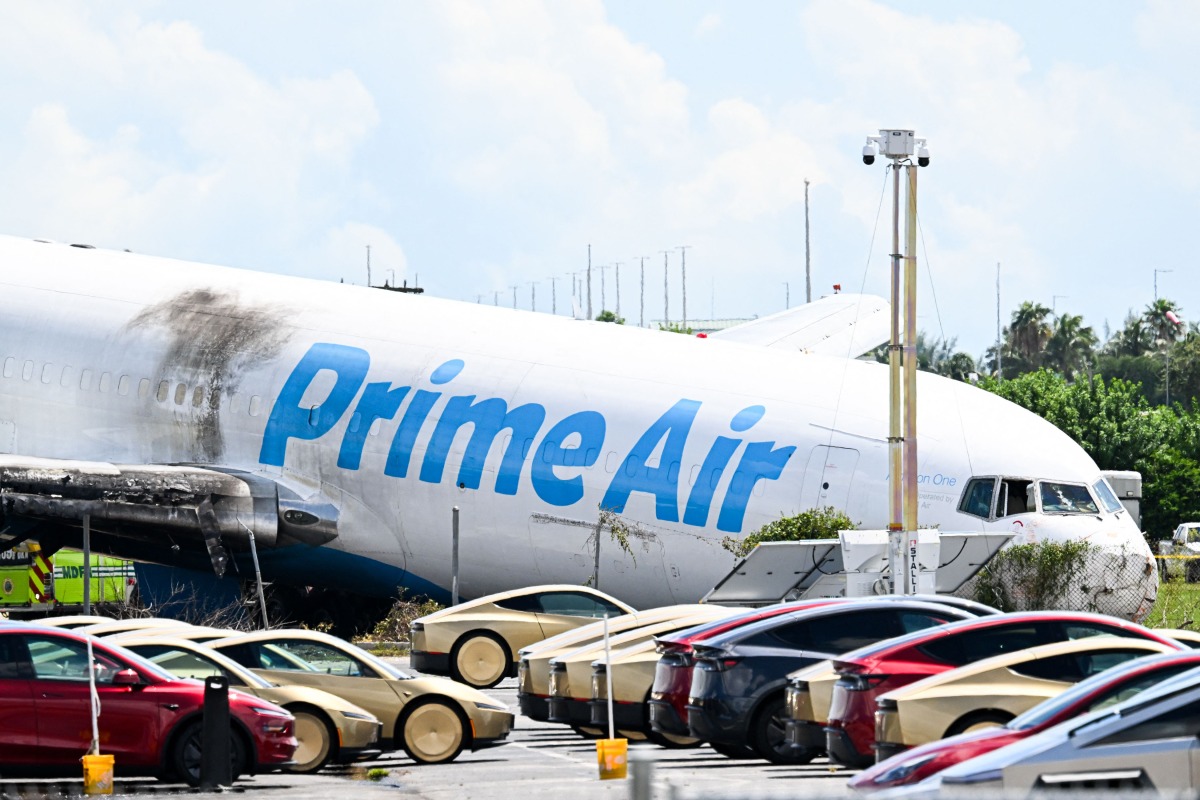 A parking lot filled with Tesla Cybercabs is seen in the foreground as the nose of a Prime Air cargo plane rests on the ground after the aircraft overran the runway and struck cars the previous day at Miami International Airport in Miami, Florida, on September 7, 2026. (Photo by CHANDAN KHANNA / AFP)