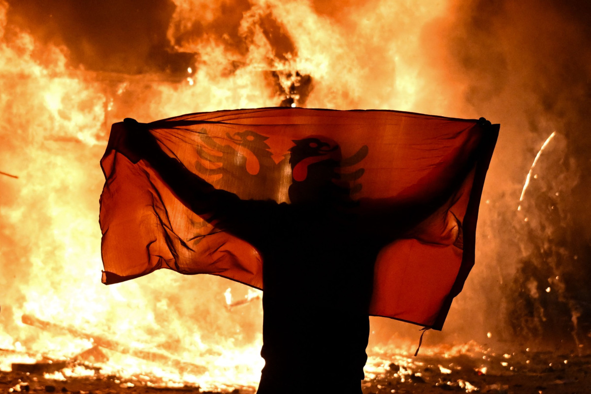 A protester holds an Albanian National flag in front of a burning kiosk during an anti-government demonstration in central Tirana on October 15, 2026, as protests continued for a third consecutive day over university conditions and demands for the resignation of Albanian Prime Minister. Photo by Armend NIMANI / AFP